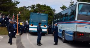 ゲート(手前左側)への搬入を阻止しようと抗議する市民らを、隊員の人垣と機動隊車両(左)でふさぎ、さらに県道上に貸し切りバスを留め置いて通行を規制した=26日正午ごろ、東村高江・米軍北部訓練場メインゲート前