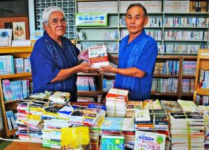 「喜屋武文庫」の書棚の前で宮里徳成教育長(右)へ寄贈本を手渡す喜屋武忠信さん=伊江村中央公民館図書室