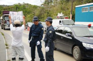 米軍北部訓練場でのヘリ着陸帯建設工事で、パトカーに先導され基地内に入る工事車両に抗議する市民=30日午前10時、東村高江の同基地N1ゲート