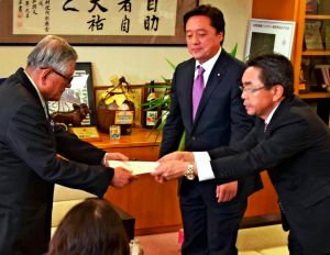 伊集盛久東村長(左)に交付決定通知書を手渡す中嶋浩一郎沖縄防衛局長(右)と若宮健嗣防衛副大臣(中央)=4日、東村役場