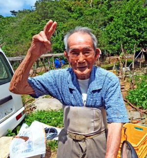 「あと2本立てれば柱は完成する。次は横棒を組み立てるよ」と話す新崎さん。後方の柱は完成間近のナーべーラー棚=名護市真喜屋