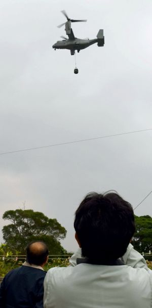 村職員らが見上げる中、物資をつり下げて旋回するオスプレイ=6日午後3時47分ごろ、宜野座村城原区内