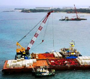 米軍普天間飛行場の移設に向けた海上の本体工事が始まり、沖縄県名護市辺野古沿岸部では海中に設置する大型コンクリート製ブロックを台船(手前)からクレーン船に移す作業が行われた。奥は米軍キャンプ・シュワブ=6日午後(小型無人機から)