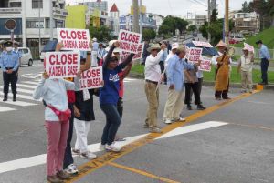 米軍嘉手納基地に向かって、基地撤去を求めるプラカードを掲げる参加者=23日午前8時22分、沖縄市山内・嘉手納基地第5ゲート前
